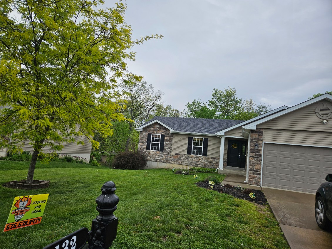 Stone and siding house with green lawn, tree, and roofing company sign