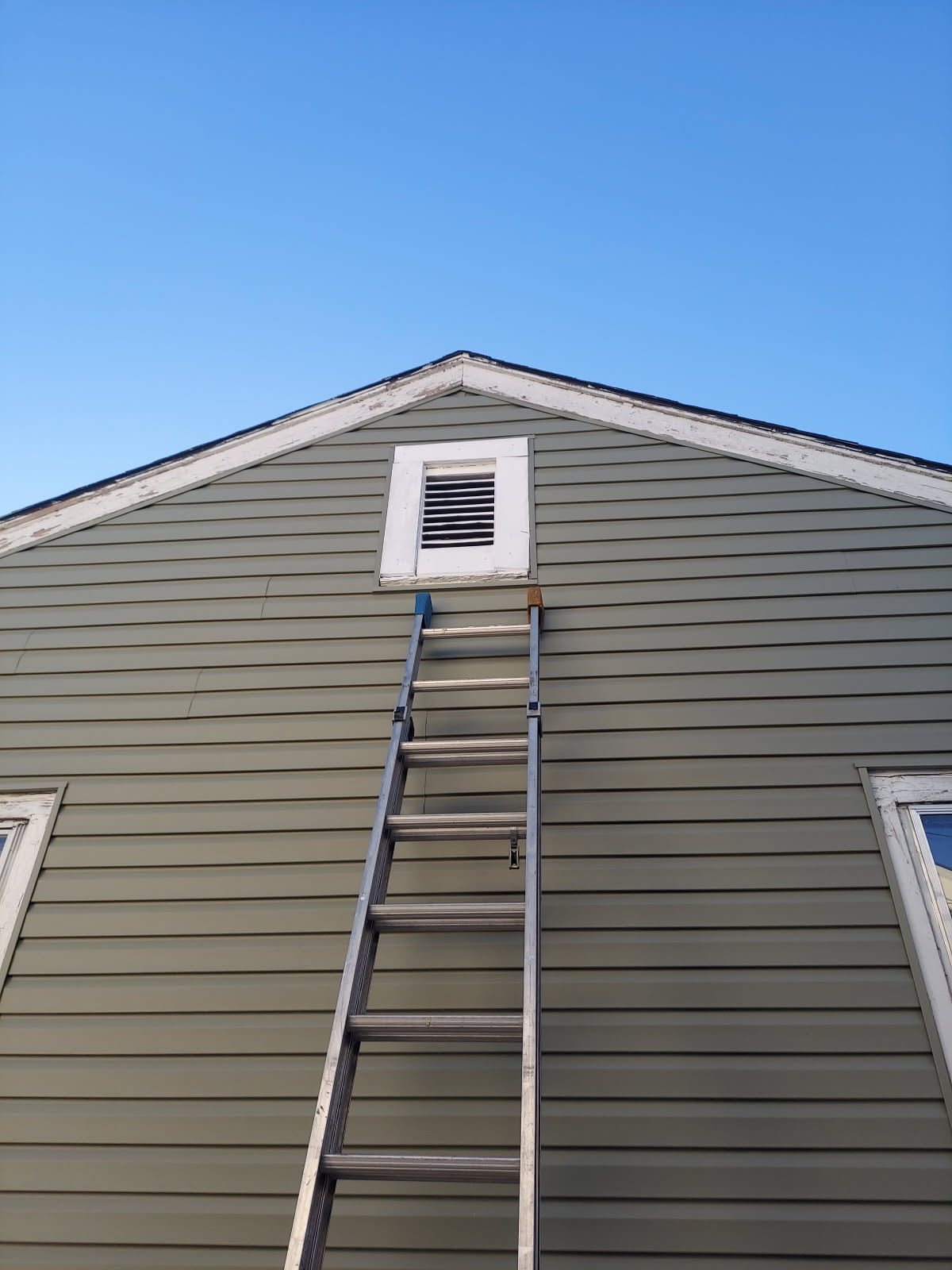 Ladder leaning against house wall leading to attic vent under blue sky